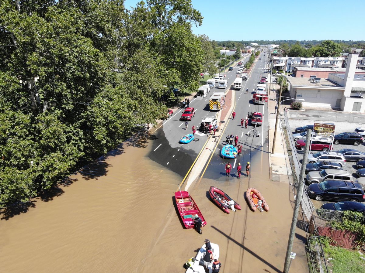 A partially flooded road with cars parked on the dry part and small boats in the flooded part.