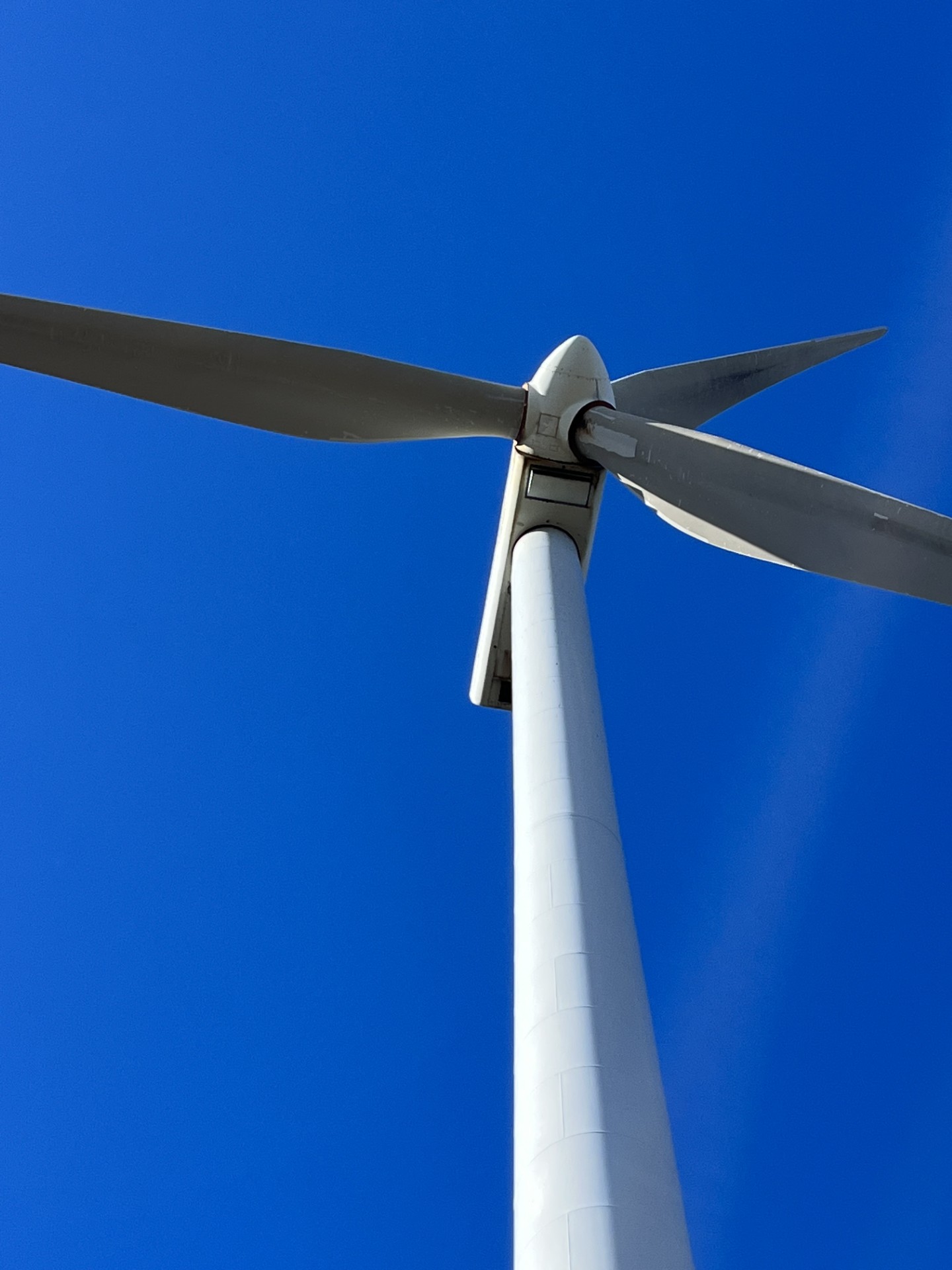 Looking up at the blades of a large wind turbine, seen against a clear blue sky.