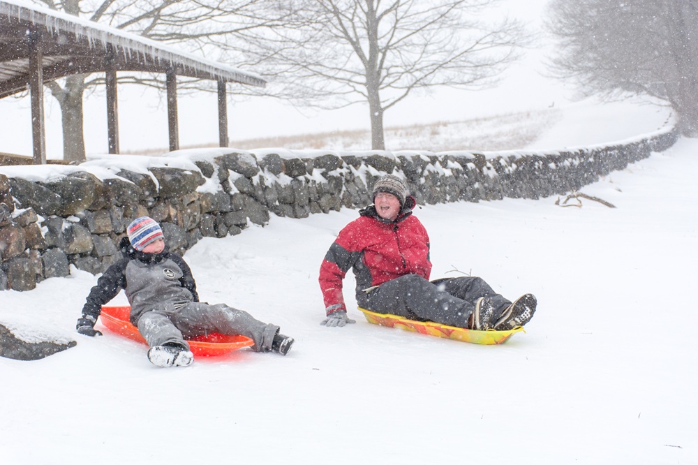 Two people sit on sleds in front of a stone fence. Snow covers the ground.