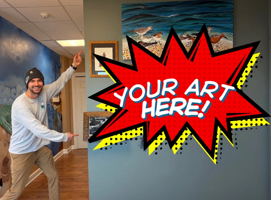 A smiling man points with both hands to a section of wall marked with a cartoon that reads "Your Art Here!"