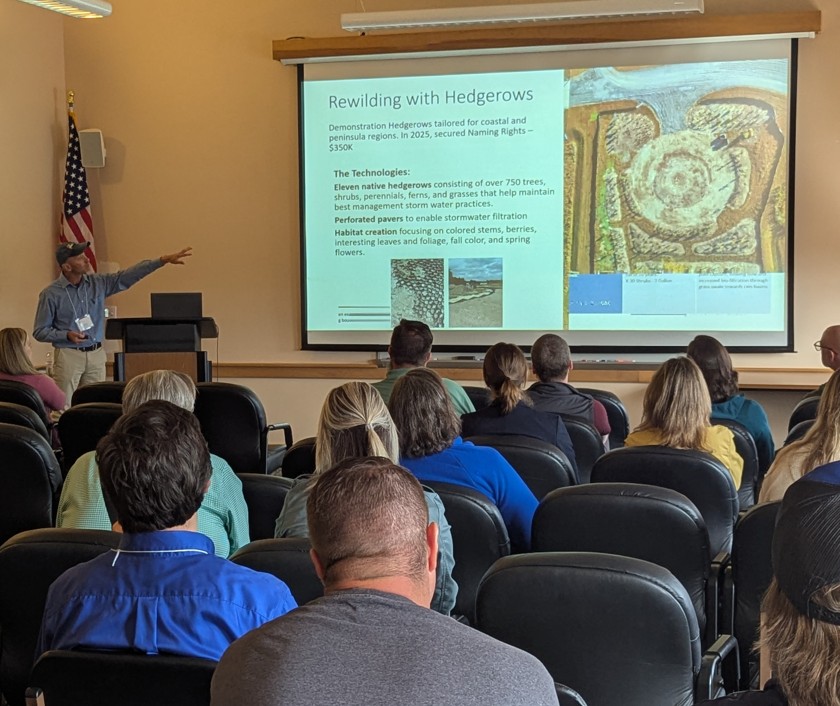 A presenter points to a screen showing plans for hedgerows in front of an audience at a meeting.