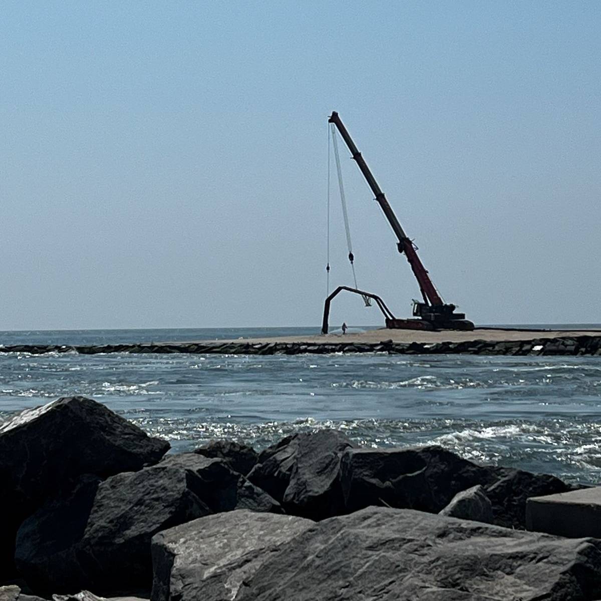 A crane seen from across a body of water, lifts a pipe that is collecting sand and water from the inter-tidal zone.