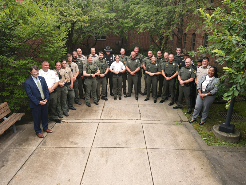 Flanked by a man and a woman in suits, about two dozen police officers in uniform stand in a semicircle in an outdoor courtyard.