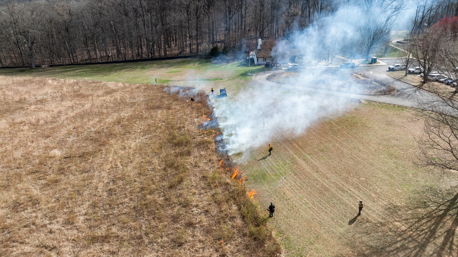 Flames and smoke rise from the edge of a field near where several individuals in protective gear stand.