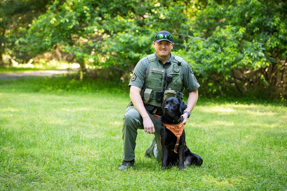 A natural resources police officer stands with a police lab dog on a leash.