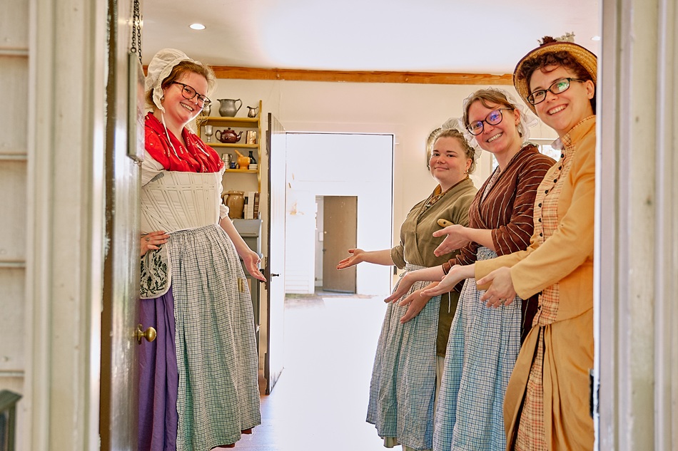 Four women dressed in colonial attire stand just inside a doorway extending greetings.