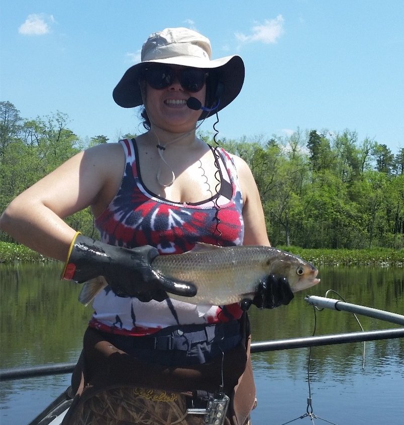 A woman holds an adult fish while standing in a boat in a river.