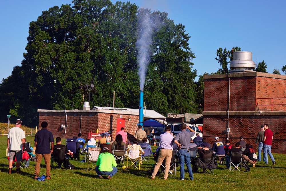 A group of people crowd around a machine as it releases a plume of smoke through a long vertical pipe.