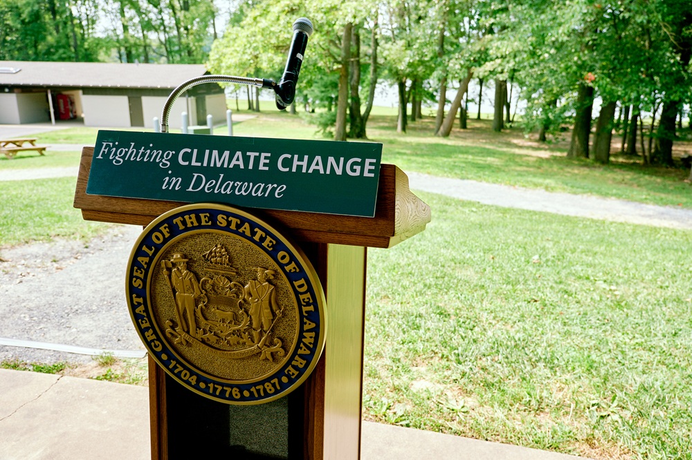 A podium with the Delaware seal and a sign about fighting climate change sits in front of trees.