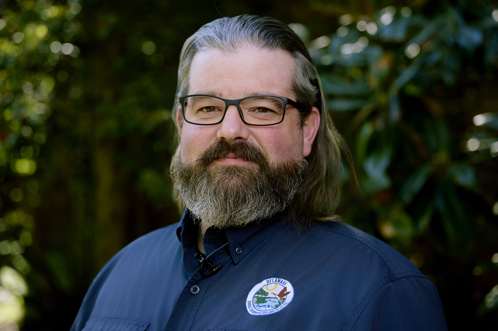 A bearded man wearing a Delaware Division of Fish and Wildlife shirt poses in front of a tree.