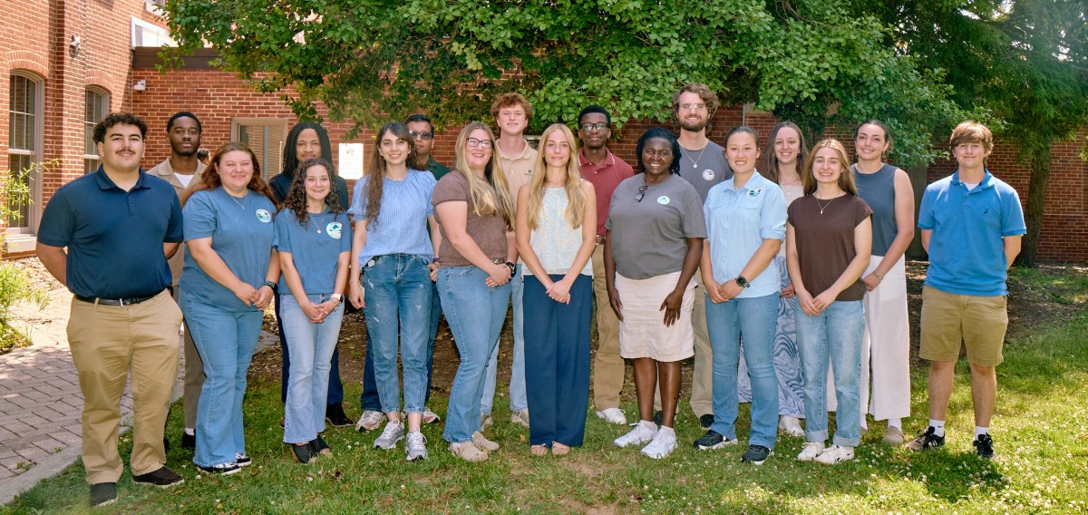 A group of 18 young adults pose in front of trees and a brick building.